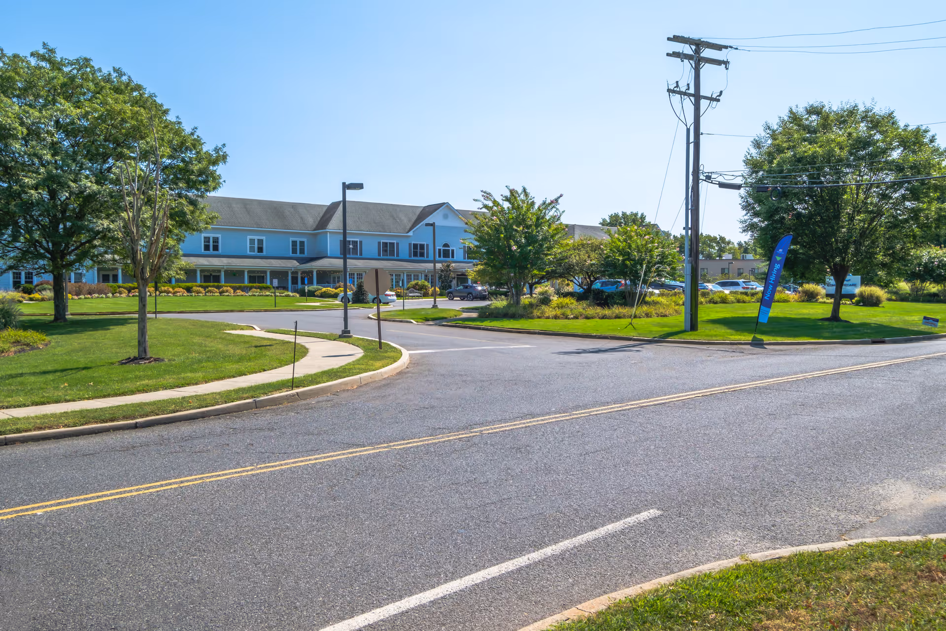 A two-story senior living building viewed from the street with a curved driveway, parking area, and landscaped lawns.