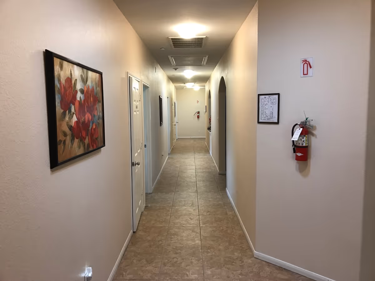 A long, narrow hallway with beige walls and tiled floor. The hallway is lit by ceiling lights and has several doors on the left side and an arched opening on the right. A framed painting of red flowers hangs on the left wall, and a fire extinguisher is mounted on the right wall near a framed floor plan.