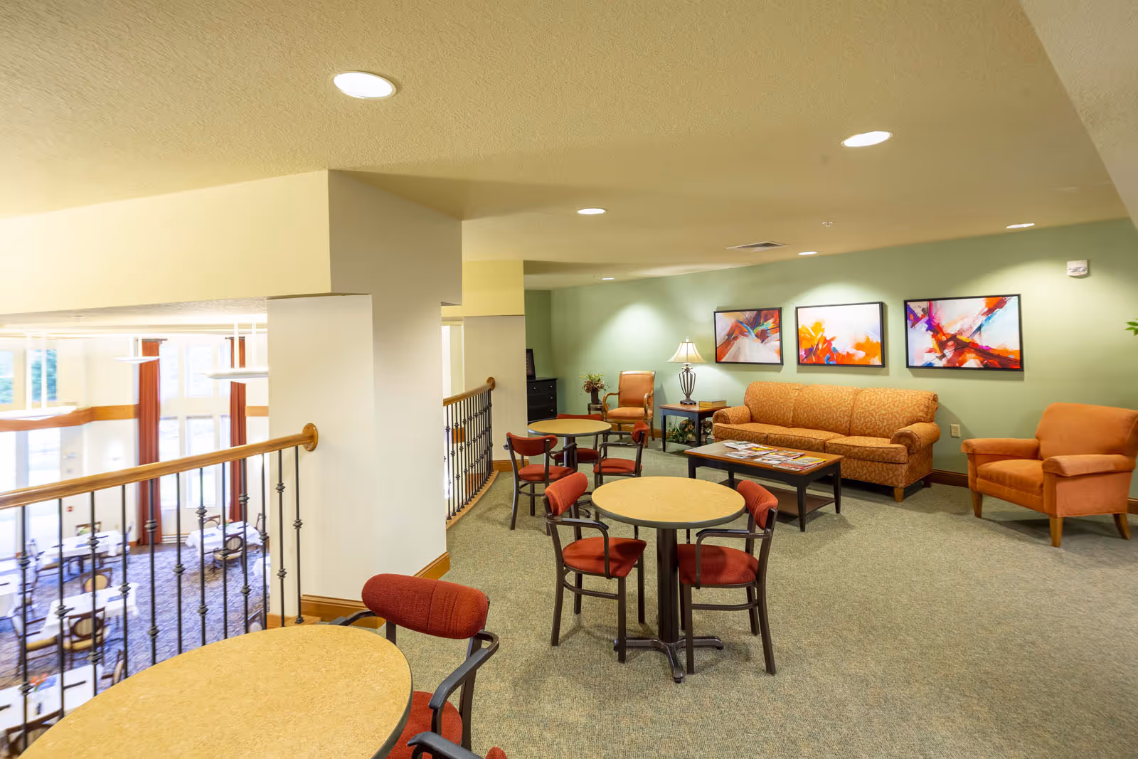An upstairs communal lounge with round tables and chairs, sofas and armchairs, and colorful abstract paintings on the wall overlooking a dining area below.