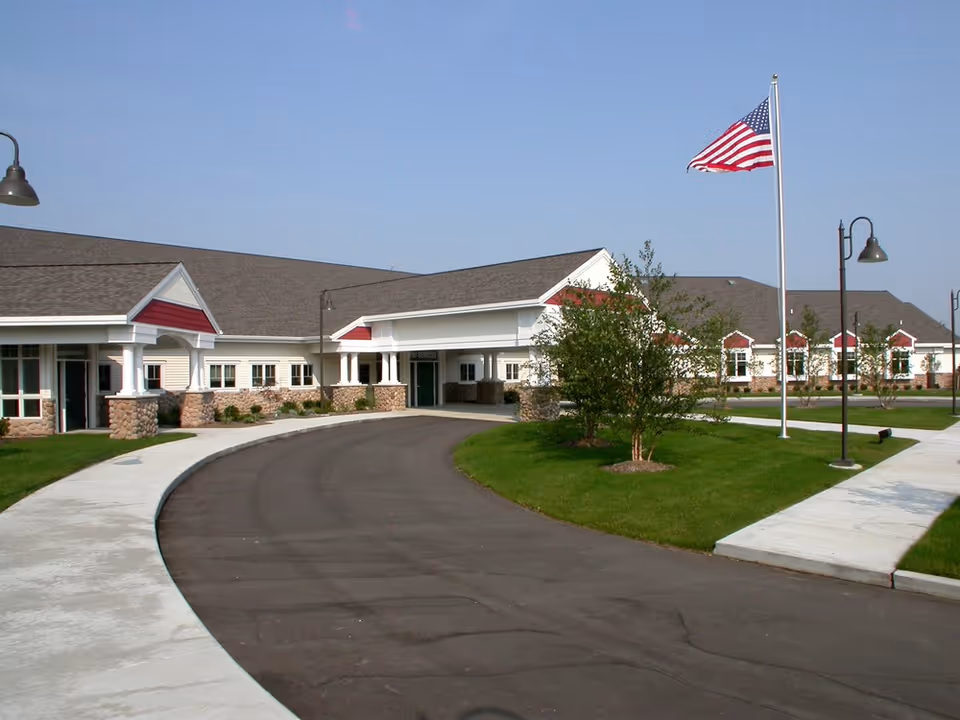 Exterior view of Care Cardinal Byron Center facility showing a single-story building with a curved driveway, green lawns, trees, street lamps, and an American flag on a flagpole against a clear blue sky.