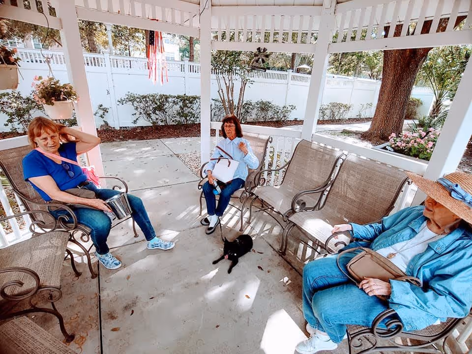 Three elderly women sitting on metal benches under a white gazebo in an outdoor garden area. One woman is wearing a blue shirt and jeans, another is in a light blue shirt and dark pants, and the third is wearing a blue jacket and a large sunhat. A small black dog is lying on the concrete floor between them. There are plants and trees surrounding the gazebo, and an American flag is hanging from the structure.