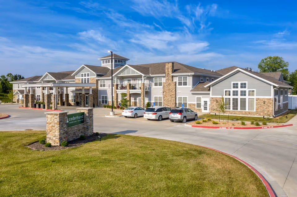 Exterior view of Cedarhurst Senior Living of Blue Springs building with a stone and gray siding facade, several parked cars, a green lawn, and a clear blue sky.