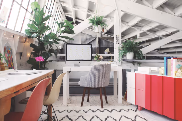Bright attic workspace with a white desk holding a computer monitor, a striped chair, various plants, a red and white cabinet, and colorful chairs along a wooden table under a slanted ceiling with exposed beams.