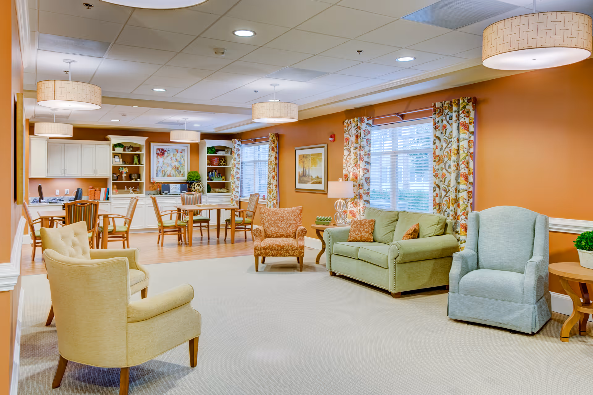 A spacious and well-lit living room area in a senior living facility with orange walls, beige carpet, and multiple seating options including a green sofa, two armchairs, and a patterned chair. The room features large windows with floral curtains, several round pendant lights hanging from the ceiling, framed artwork on the walls, and a built-in white shelving unit with decorative items and a desk area with chairs.