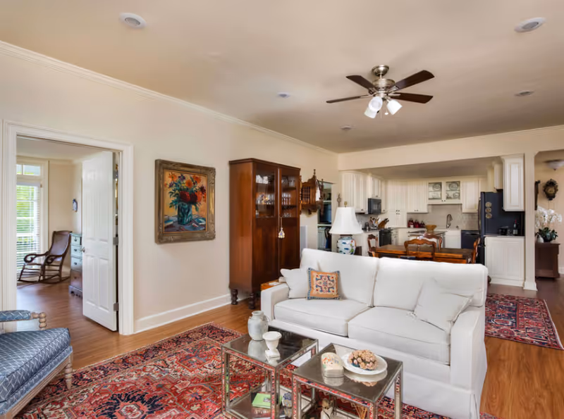A cozy living room area with a white sofa, glass coffee tables, and a colorful patterned rug. The room opens into a kitchen and dining area with white cabinetry and wooden furniture. A ceiling fan with lights is mounted on the ceiling, and a framed floral painting hangs on the wall. There is a doorway leading to another room with a rocking chair and dresser visible.
