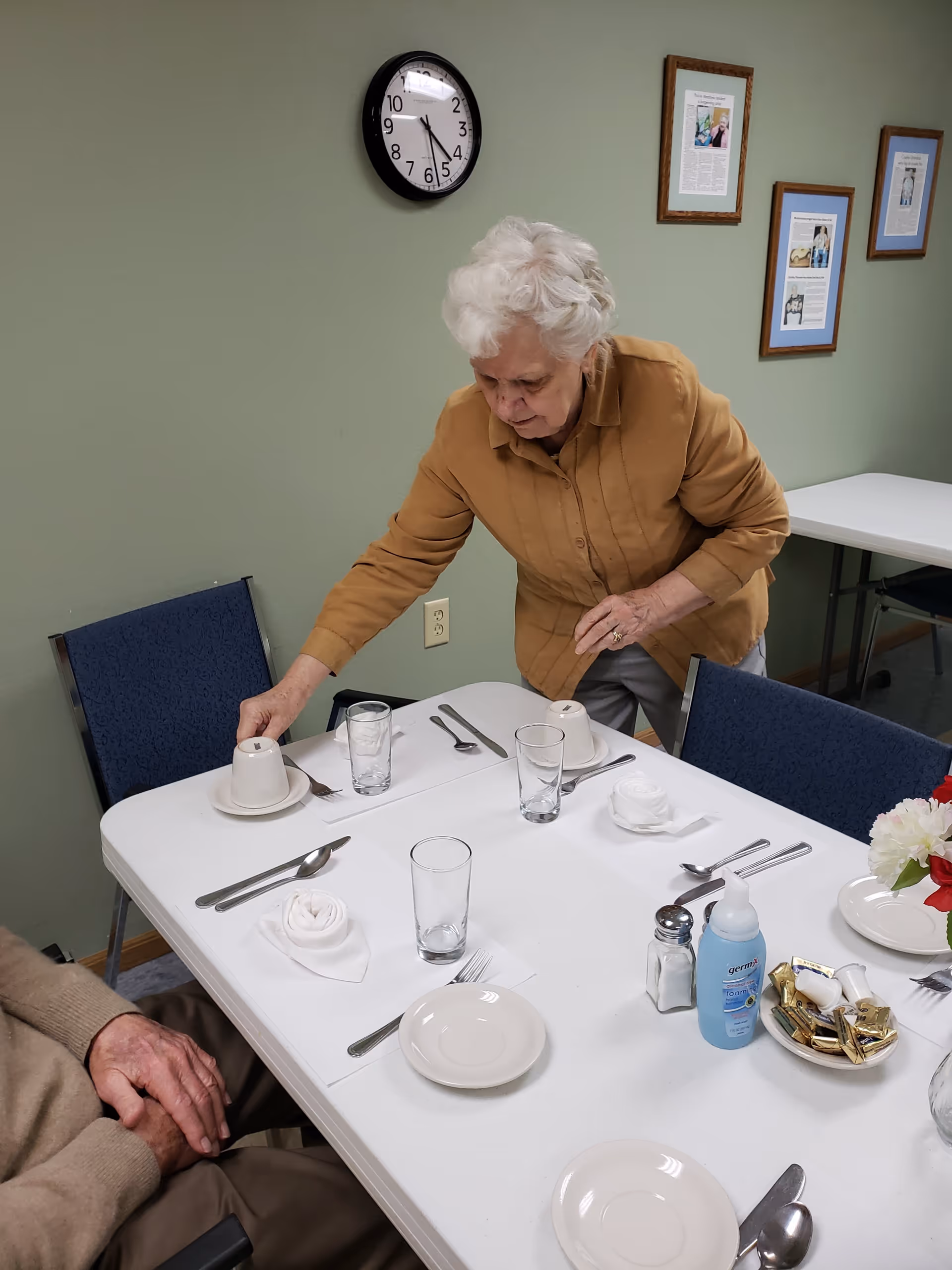 An elderly woman with white hair wearing a brown shirt is setting a dining table with white plates, glasses, utensils, and napkins folded into rose shapes. A man is seated at the table. The room has green walls with framed articles, a clock showing 5:27, and blue chairs around the table. There is a bottle of Germ-X hand sanitizer, a salt shaker, and a bowl of wrapped candies on the table.