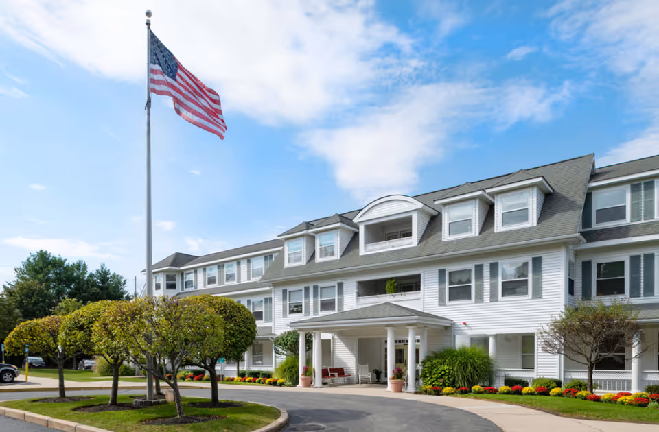 Exterior view of a large white senior living facility building with multiple windows and a covered entrance. There is a circular driveway with neatly trimmed bushes and colorful flowers along the walkway. An American flag on a tall flagpole is prominently displayed in front of the building under a partly cloudy sky.