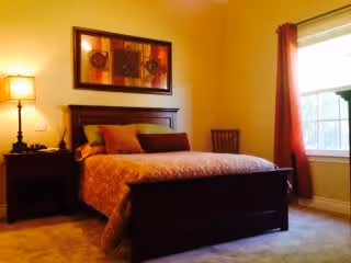 A warmly lit bedroom featuring a wooden bed with a patterned bedspread and multiple pillows. There is a nightstand with a lamp on the left side of the bed, a framed artwork hanging above the headboard, and a window with red curtains on the right side letting in natural light.