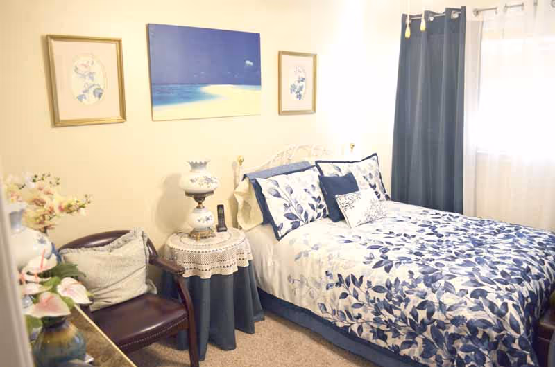 A cozy bedroom with a bed dressed in blue and white floral bedding, a small round table with a lace tablecloth and a decorative lamp, a dark wooden chair with cushions, floral artwork on the walls, and a window with dark blue curtains letting in natural light.