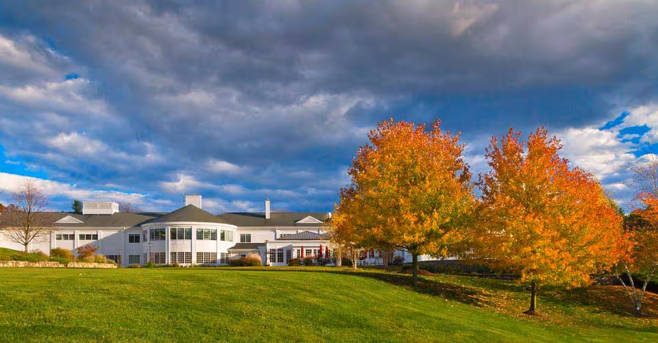 A large white building with multiple windows and a rounded section, set against a dramatic cloudy sky. In the foreground, there is a green grassy lawn and several trees with autumn-colored leaves in shades of orange and yellow.