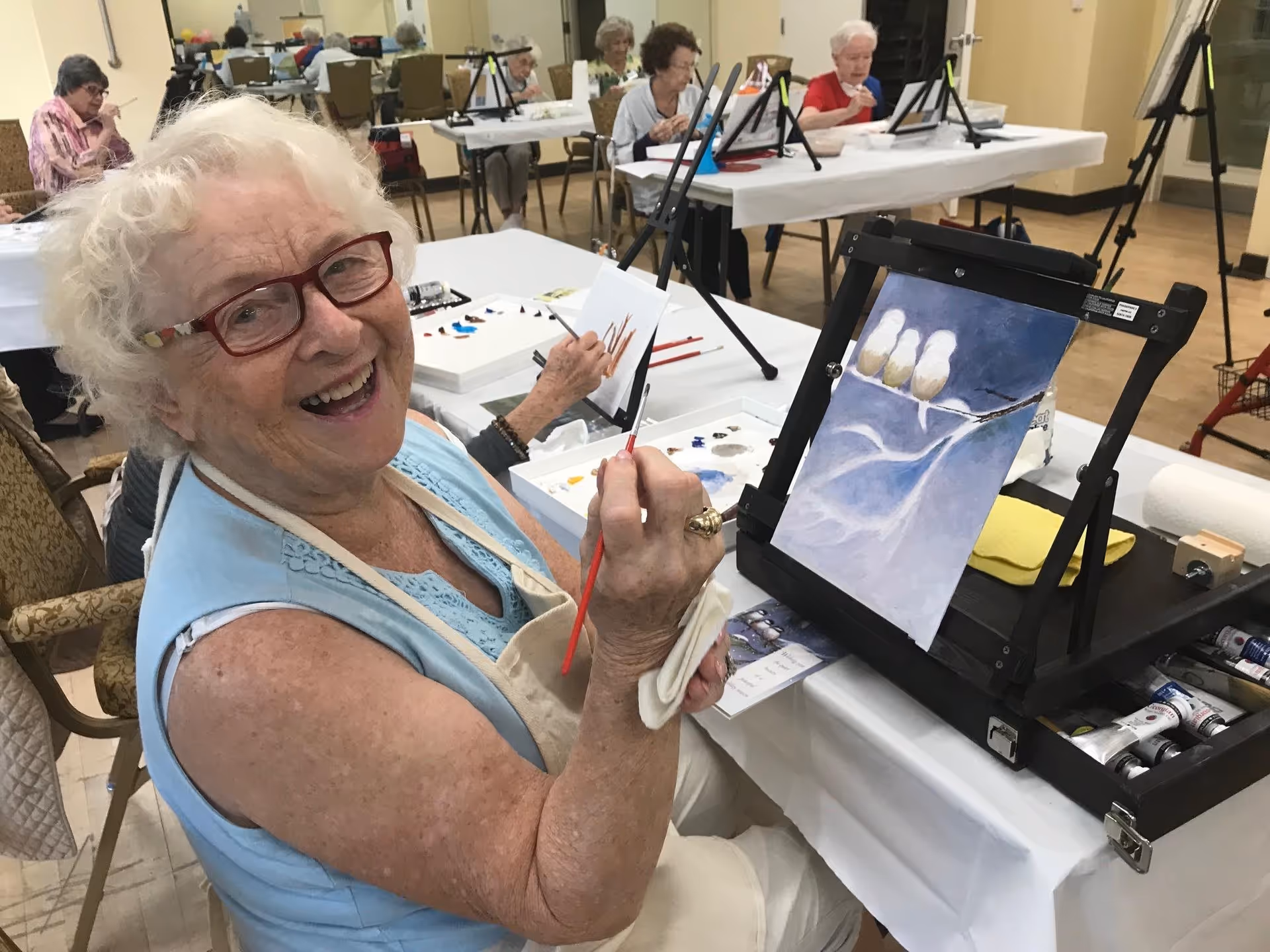 An elderly woman with white hair and red glasses smiling while painting on a canvas in an art class. Several other elderly individuals are also painting at tables in the background in a well-lit room.