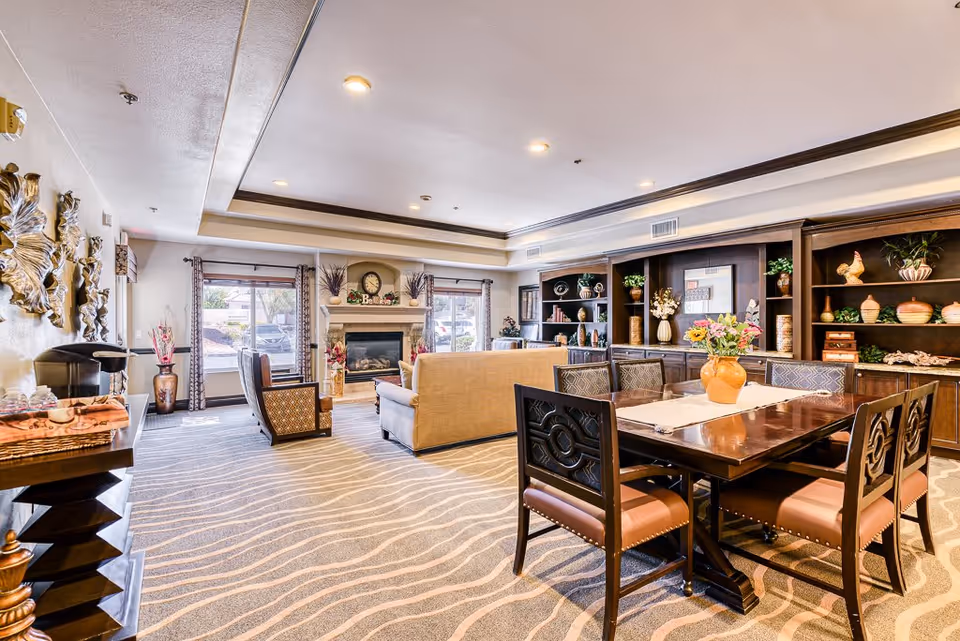 A spacious and well-lit living room area in a senior living facility with a beige sofa, two armchairs, a wooden dining table with six chairs, decorative shelves with plants and pottery, a fireplace with a clock and floral decorations, and large windows with curtains allowing natural light.