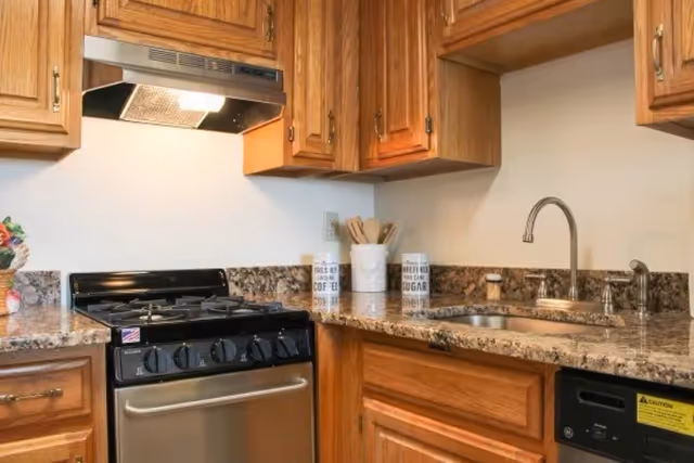 A kitchen area with wooden cabinets, a stainless steel gas stove with an overhead vent, granite countertops, a stainless steel sink with a curved faucet, and kitchen utensils in containers on the counter.