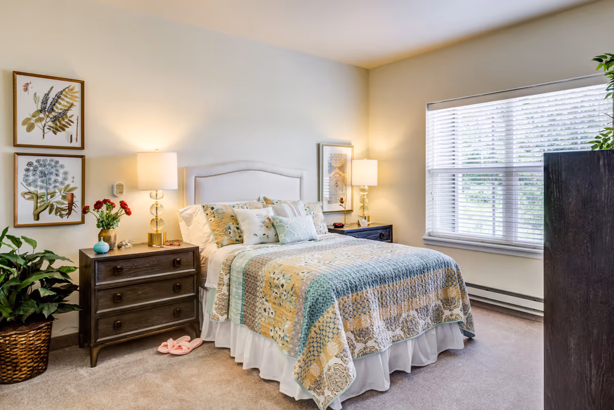 A cozy bedroom with a neatly made bed featuring a patterned quilt and multiple pillows. There are two bedside tables with lamps on each side of the bed. On the left side, there are two framed botanical prints on the wall above the bedside table, which also holds a vase with red flowers and some small decorative items. A large window with white blinds lets in natural light on the right side of the room. A potted plant is placed on the floor near the left bedside table, and a dark wooden dresser is partially visible on the right side of the image.