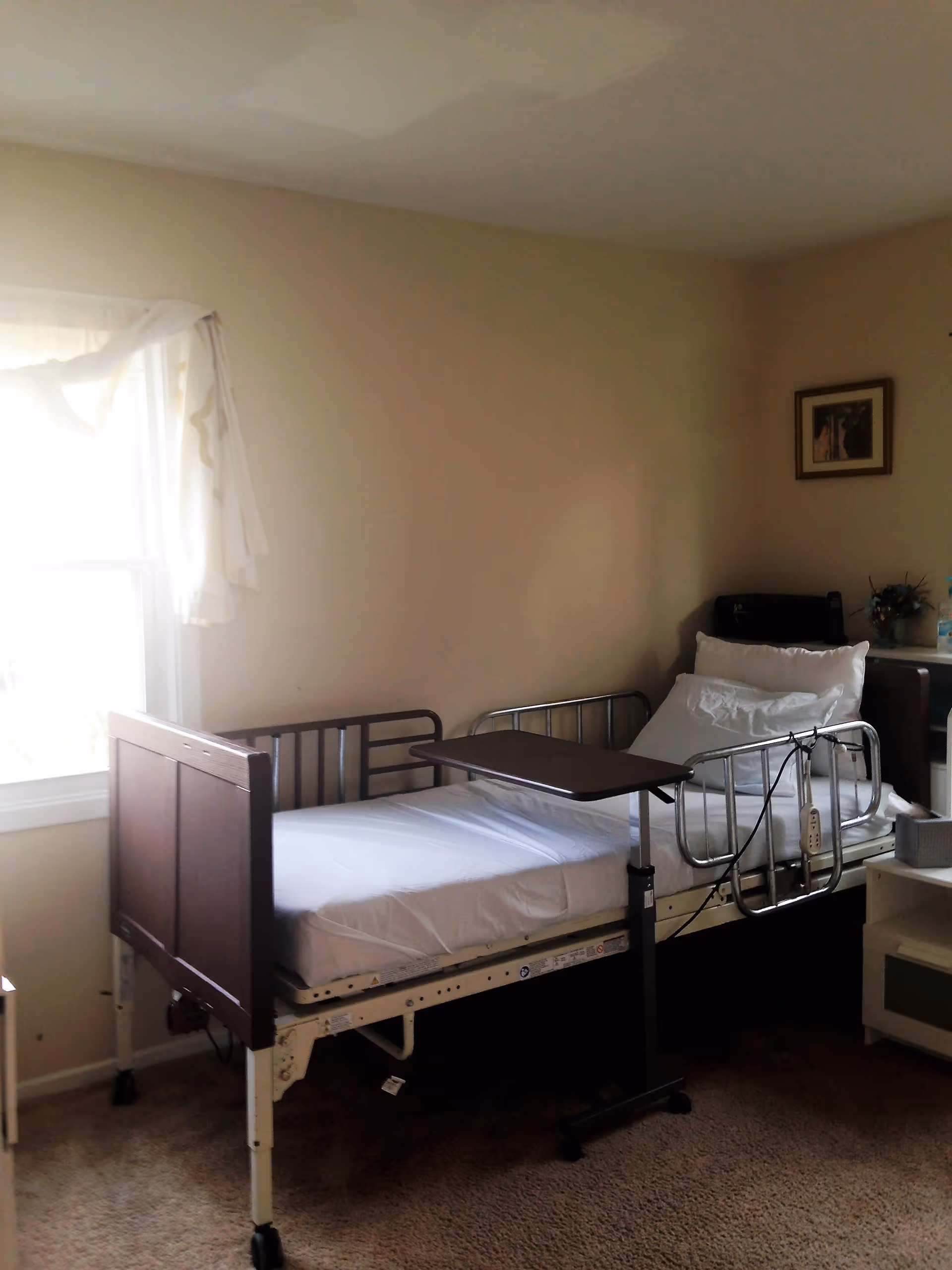 A simple room with a hospital-style bed featuring metal side rails and a wooden headboard. The bed has white sheets and pillows, and a movable overbed table is positioned above it. There is a window with sheer white curtains on the left side, and a small table with a few items including a flower arrangement and a framed picture on the wall to the right.