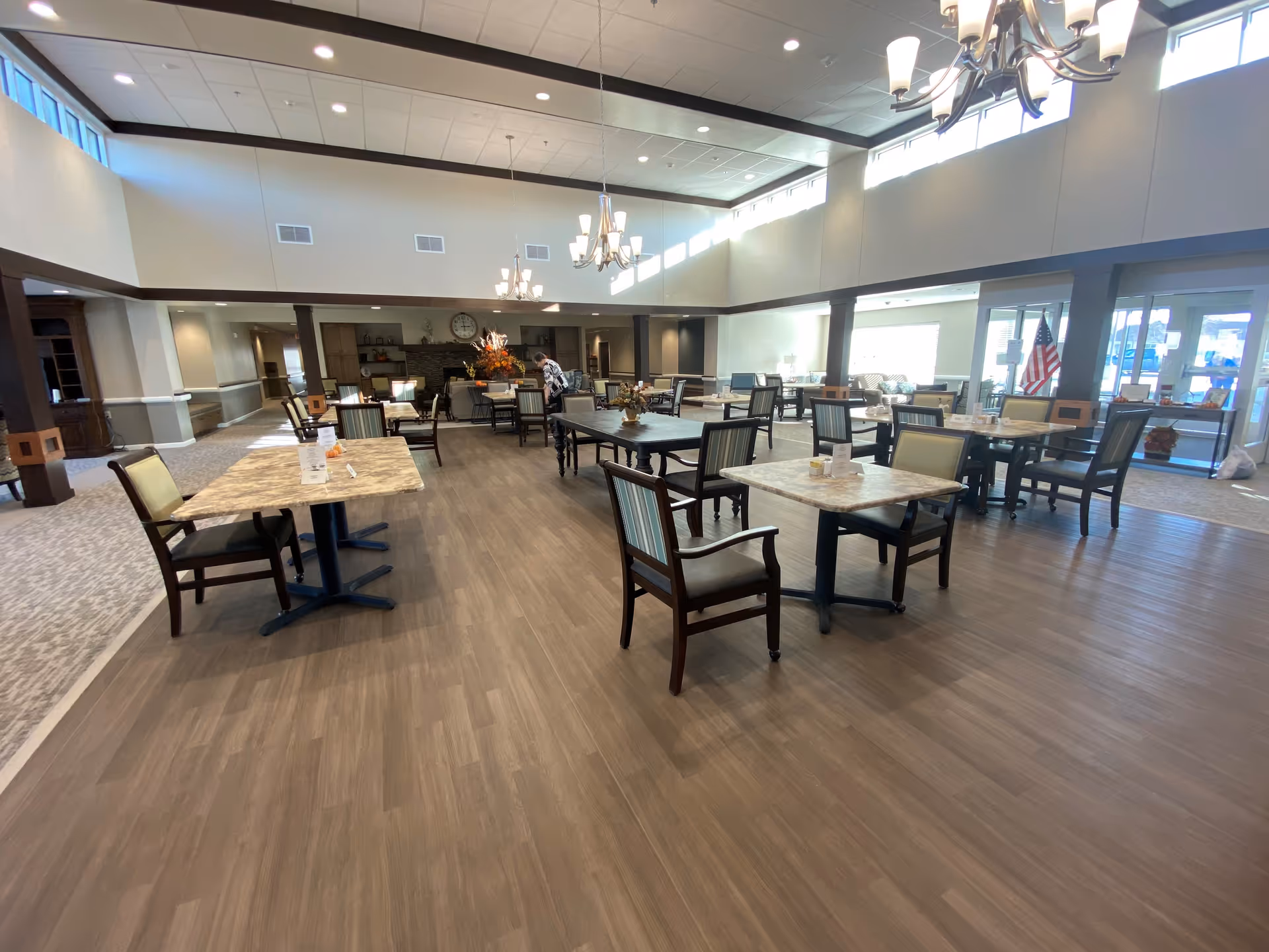 A spacious and well-lit dining area in Prairie Village Retirement Center with multiple tables and chairs arranged neatly. The room features high ceilings with chandeliers, large windows allowing natural light, and a cozy fireplace in the background. There is an American flag near the entrance and a person standing near the fireplace.