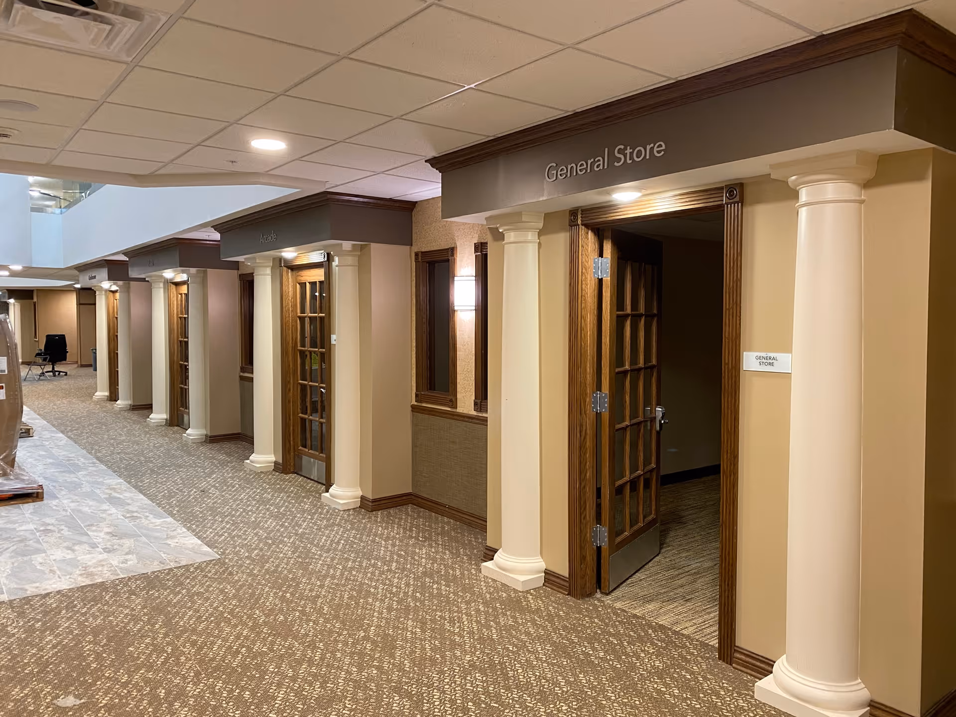 Interior hallway of Parkers Bend Retirement Community featuring multiple doorways with wooden frames and white columns. One doorway is labeled 'General Store' with an open door. The hallway has beige walls, carpeted floors, and ceiling lights.