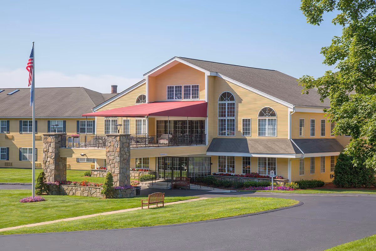 Exterior view of a large yellow senior living facility building with multiple windows and a red awning over the entrance. The building is surrounded by well-maintained green lawns, flower beds, a flagpole with an American flag, and a wooden bench near the driveway.