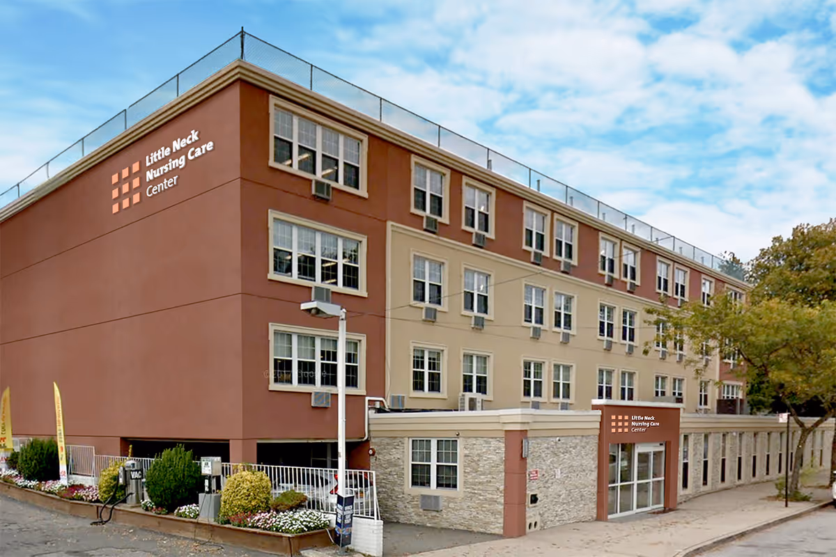 Exterior view of Little Neck Nursing Care Center, a multi-story building with beige and brown walls, multiple windows, and a main entrance with glass doors. There are trees and a sidewalk in front of the building under a partly cloudy sky.