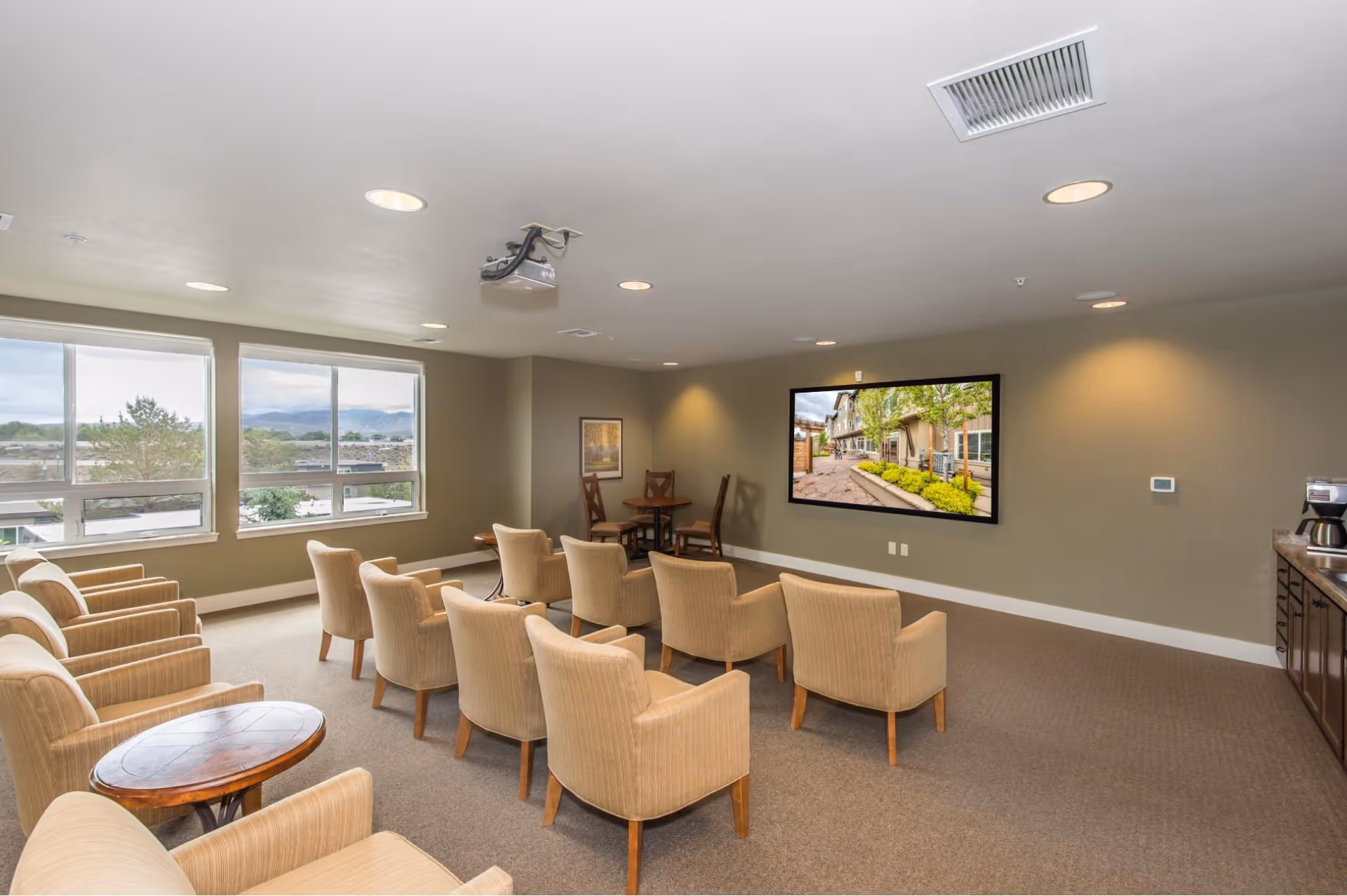 A cozy community room with beige armchairs arranged in rows facing a large wall-mounted flat screen TV displaying an outdoor scene. The room has large windows letting in natural light, a small round table with three chairs in the corner, recessed ceiling lights, and a countertop with a coffee maker on the right side.