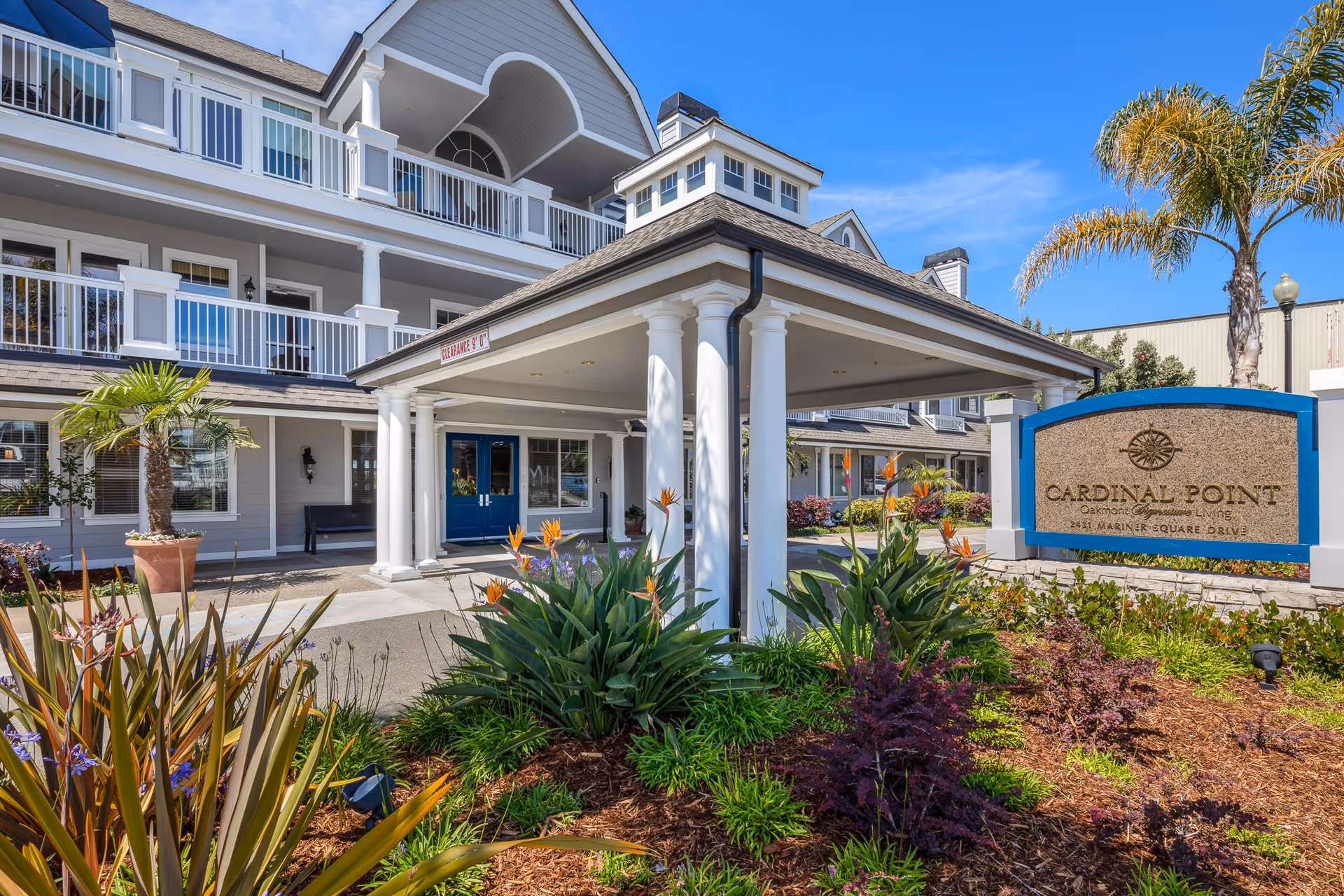 Exterior view of Cardinal Point senior living facility showing the entrance with white columns and a covered driveway. The building has multiple balconies and large windows. There are landscaped plants and flowers in the foreground, along with a sign that reads 'Cardinal Point Oakmont Senior Living 2851 Mariner Square Drive'. A clear blue sky is visible above.