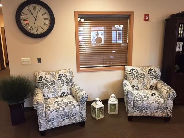 Two floral patterned armchairs placed side by side in a room with beige walls and carpet. Between the chairs on the floor are two white lanterns with green candles inside. Above the chairs is a window with wooden blinds, and to the left is a large round wall clock showing the time as 11:55. A tall potted plant is on the left side, and a dark wooden cabinet is partially visible on the right.