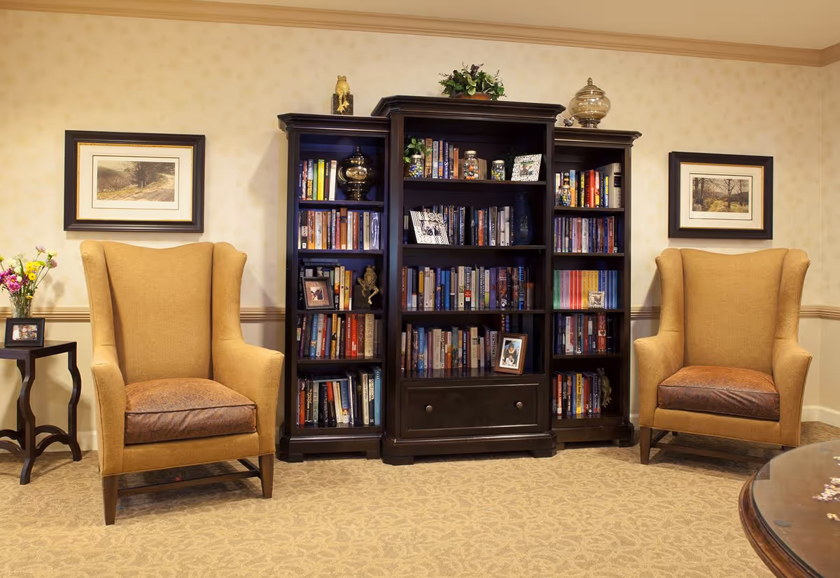 A cozy living room area with two mustard yellow wingback chairs on either side of a dark wooden bookshelf filled with books and decorative items. There are framed pictures on the wall above each chair and a small side table with a vase of flowers and a framed photo next to the left chair. The room has beige patterned carpet and wallpaper with a subtle design.