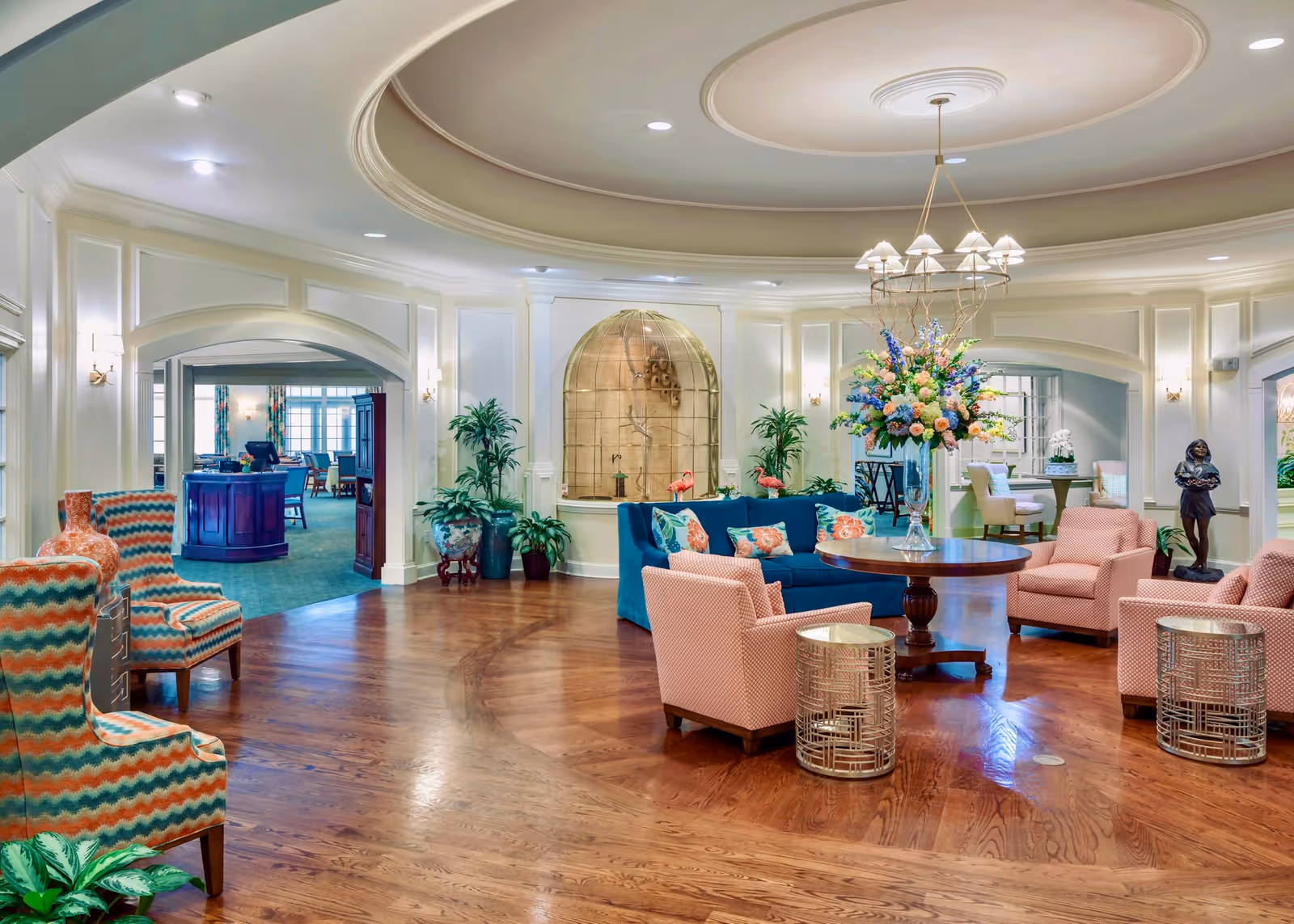 A spacious and elegant living room area with polished wooden floors, a round wooden table with a large floral arrangement, several upholstered chairs in pink and blue with patterned cushions, potted plants, and a decorative wall niche with a sculpture. The room has a coffered ceiling with recessed lighting and a chandelier, and there are archways leading to other rooms in the background.