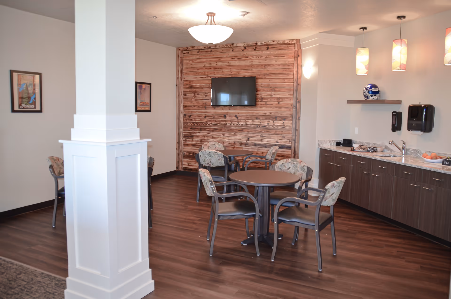 A senior living facility common area with round tables and chairs, a wooden accent wall with a mounted flat-screen TV, a kitchenette with a sink, countertop, and cabinets, pendant lights hanging from the ceiling, and framed pictures on the walls.