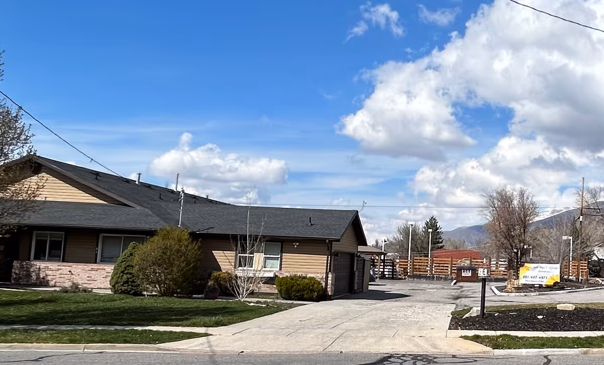 Exterior view of a single-story building with a sloped roof, surrounded by green grass, bushes, and trees under a partly cloudy blue sky. There is a driveway leading to the building and a sign near the entrance.