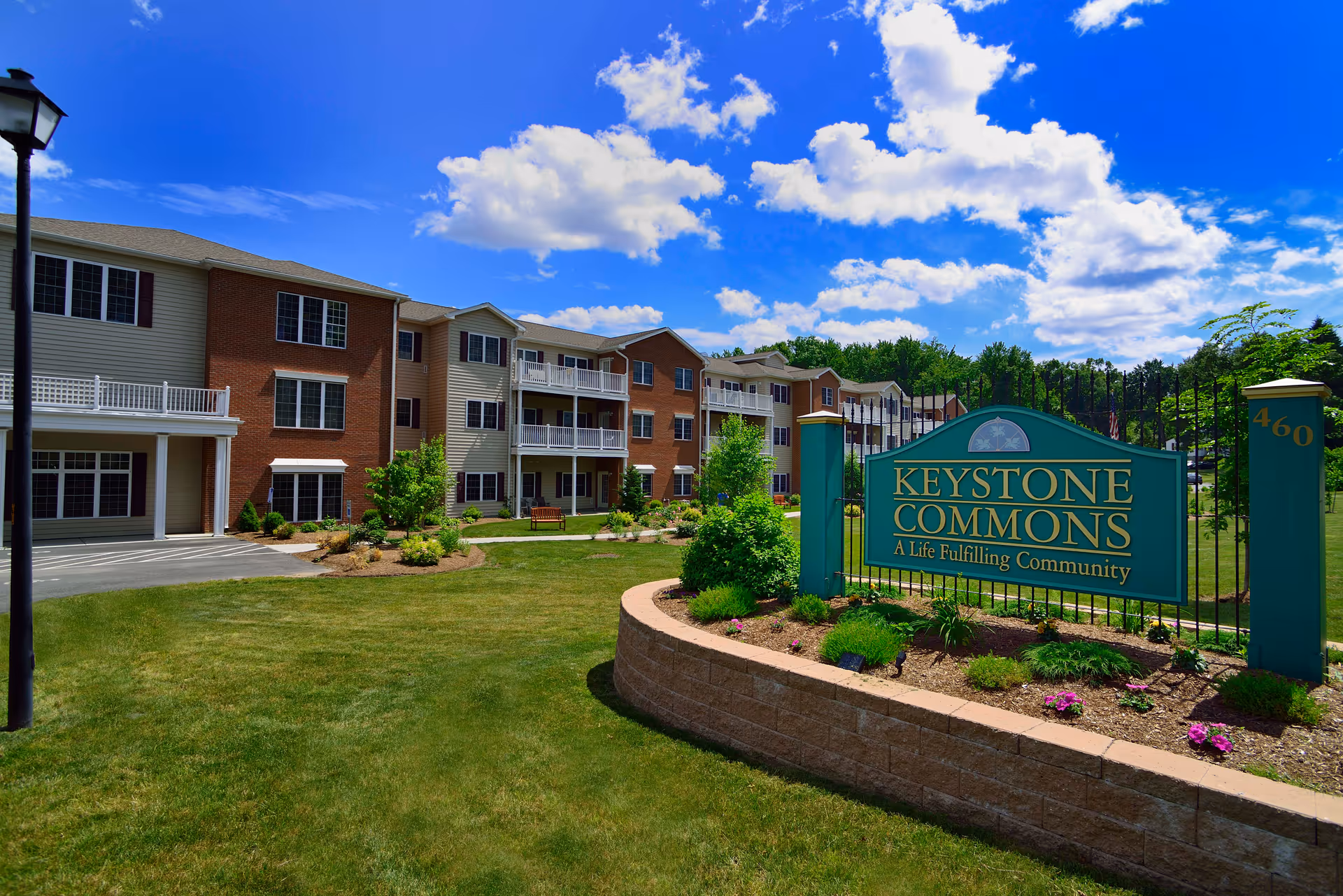 Exterior view of Keystone Commons, a senior living community, showing a large green sign with the community name and slogan 'A Life Fulfilling Community' in front of a well-maintained lawn and a multi-story residential building under a partly cloudy blue sky.