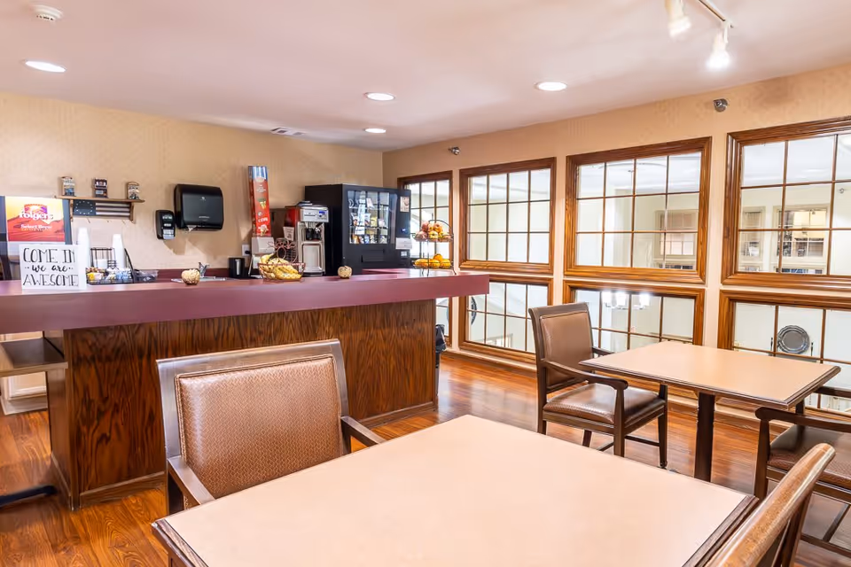 A small dining area with tables and chairs in front of a counter stocked with coffee supplies, a vending machine, and a tiered fruit stand. Large windows with wooden frames overlook an interior space below. The room has wooden flooring and beige walls.