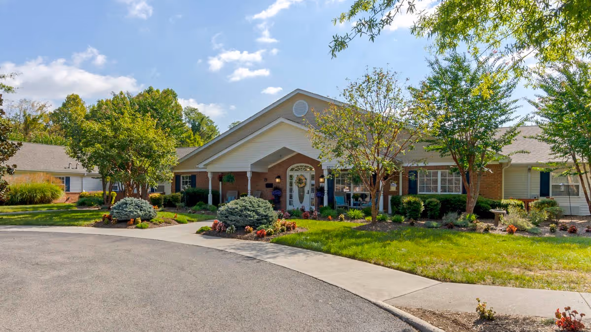 Front entrance of a single-story senior living building with a covered porch, landscaped lawns, and trees under a blue sky.