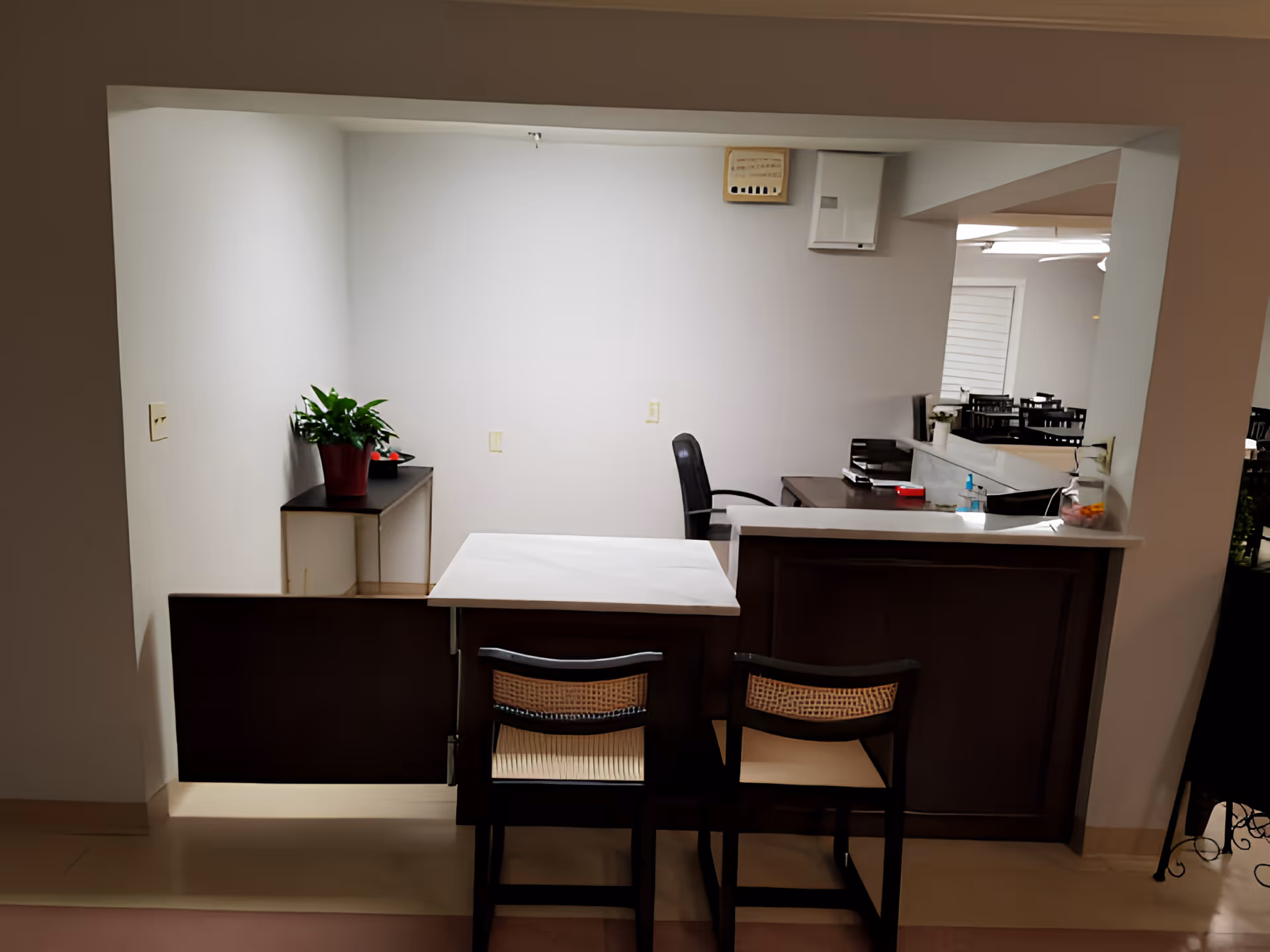 Interior reception area with a countertop and two chairs, an office desk and chair behind it, a small table with a potted plant, and dining tables visible in the background.