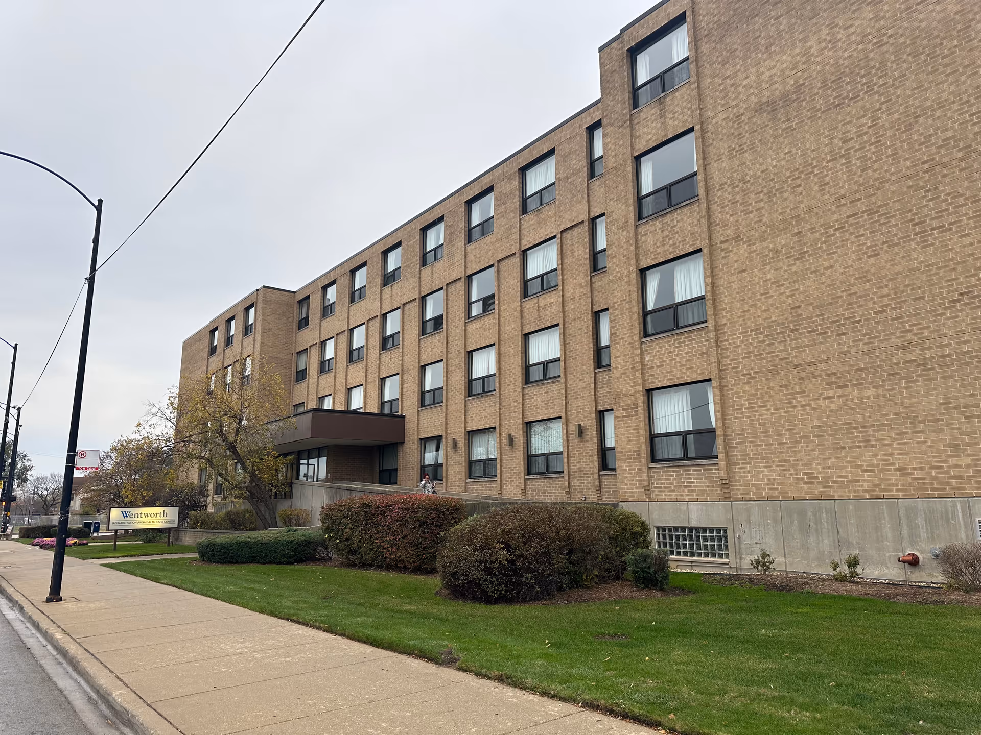 Exterior view of a multi-story brick building with numerous windows, a sidewalk, green lawn, bushes, and a sign that reads Wentworth Rehabilitation and Health Care Center under an overcast sky.