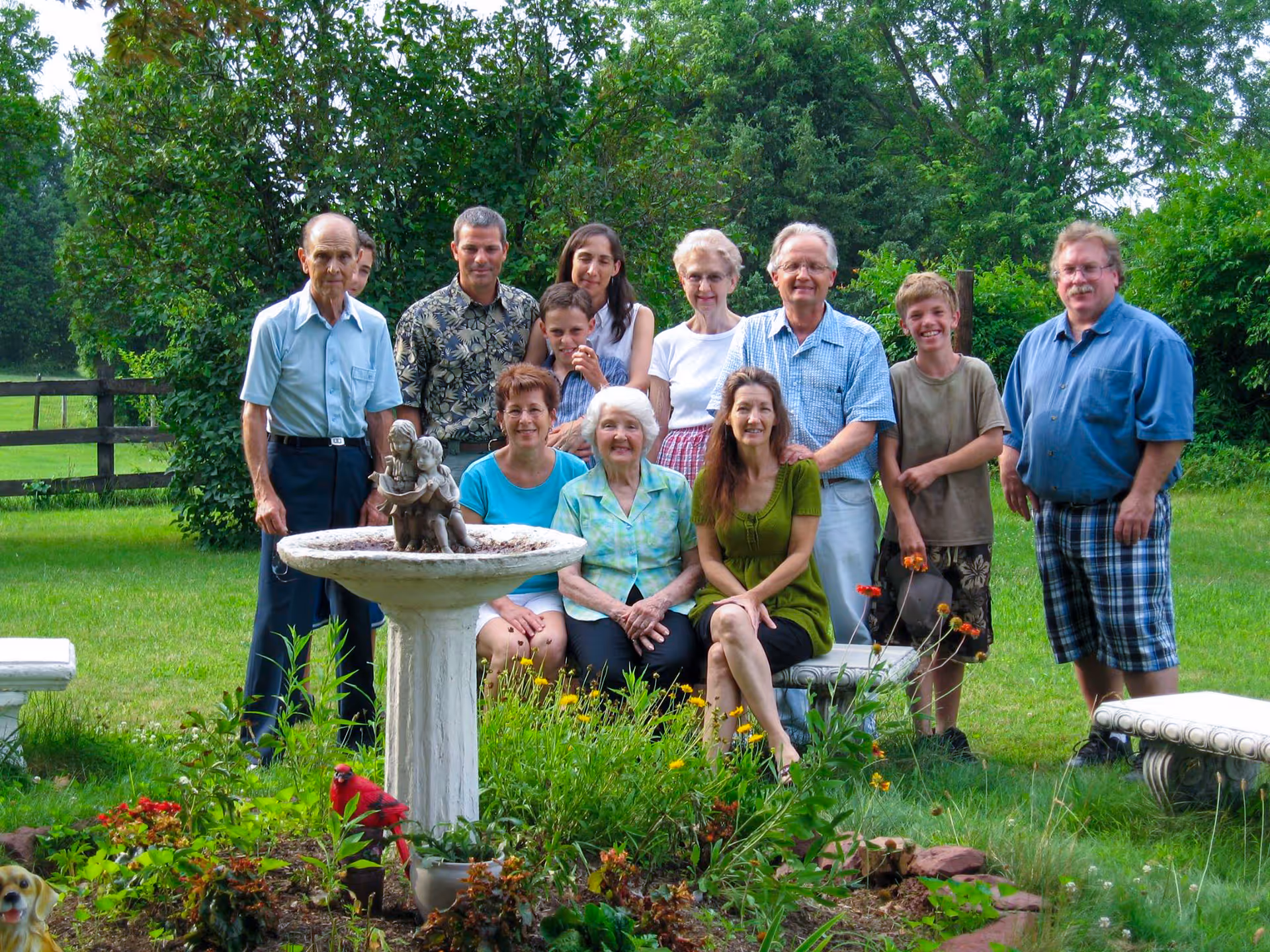 A group of eleven people of various ages posing together outdoors in a garden area with green grass, trees, and bushes. In the foreground, there is a white birdbath with a small statue on top and colorful flowers around it. The group includes older adults, middle-aged adults, and children, all smiling and standing or sitting near the birdbath.