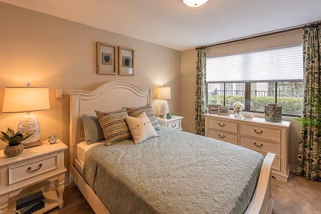 Well-lit bedroom with a made bed, matching nightstands and lamps, a dresser under a window, and patterned curtains.