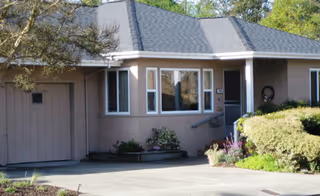 A single-story beige senior living facility front with driveway, windows, and shrubs.