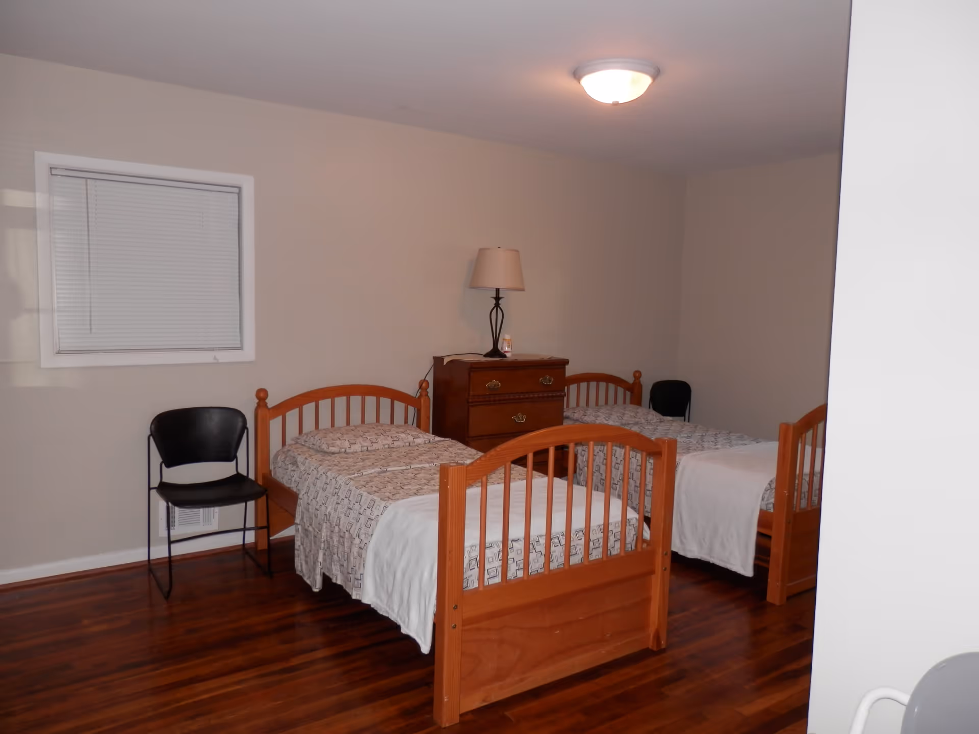 Two wooden twin beds in a sparsely furnished bedroom with a dresser, lamp, and chairs on hardwood floors.