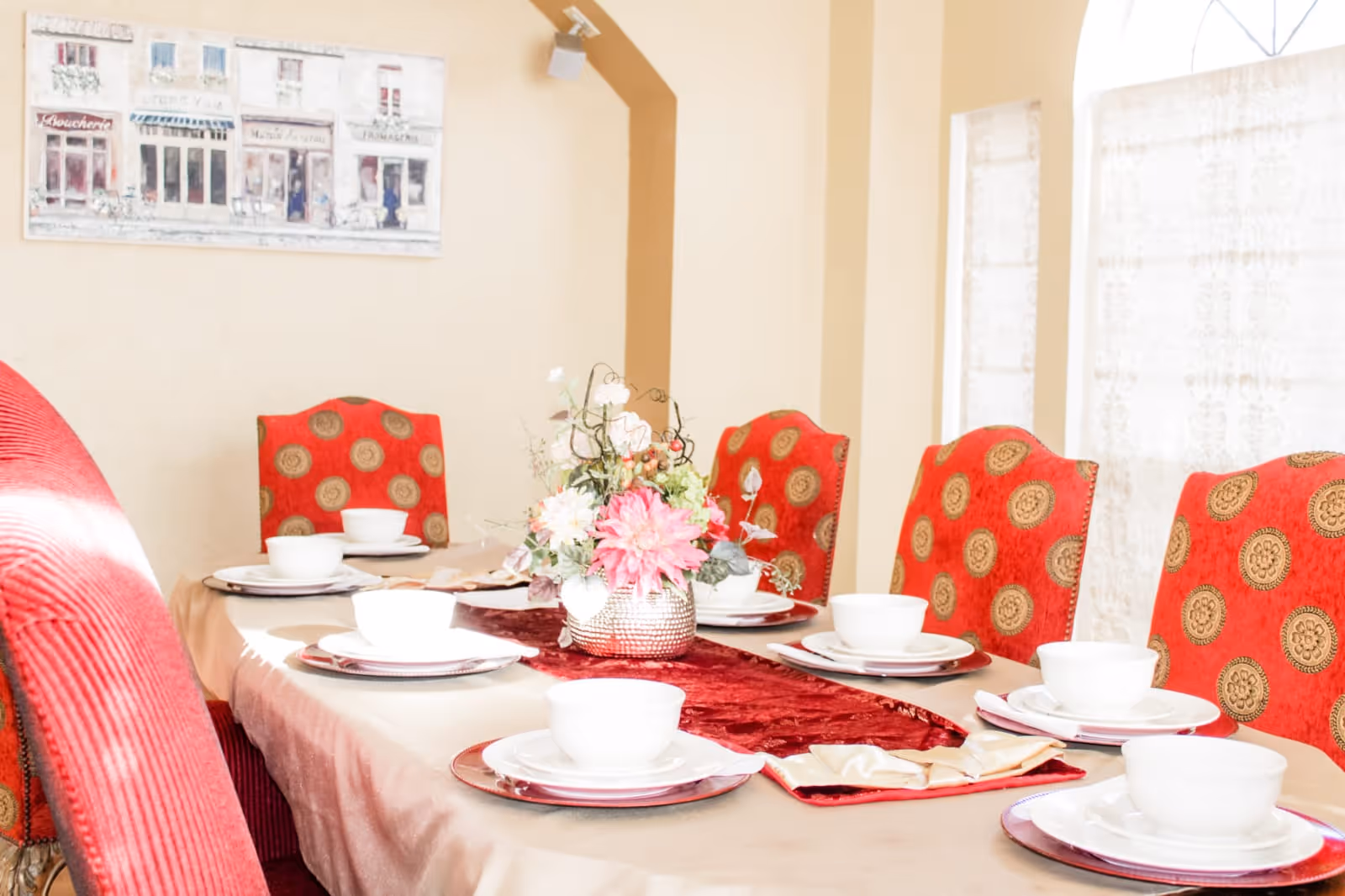 A dining table set for six with white bowls and plates on red chargers, a beige tablecloth with a red runner, and a floral centerpiece. The chairs are upholstered in red fabric with a gold circular pattern. The room has beige walls and a window with sheer white curtains.