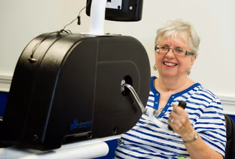 An elderly woman with short gray hair and glasses is smiling while using a hand-crank exercise machine indoors. She is wearing a blue and white striped shirt.