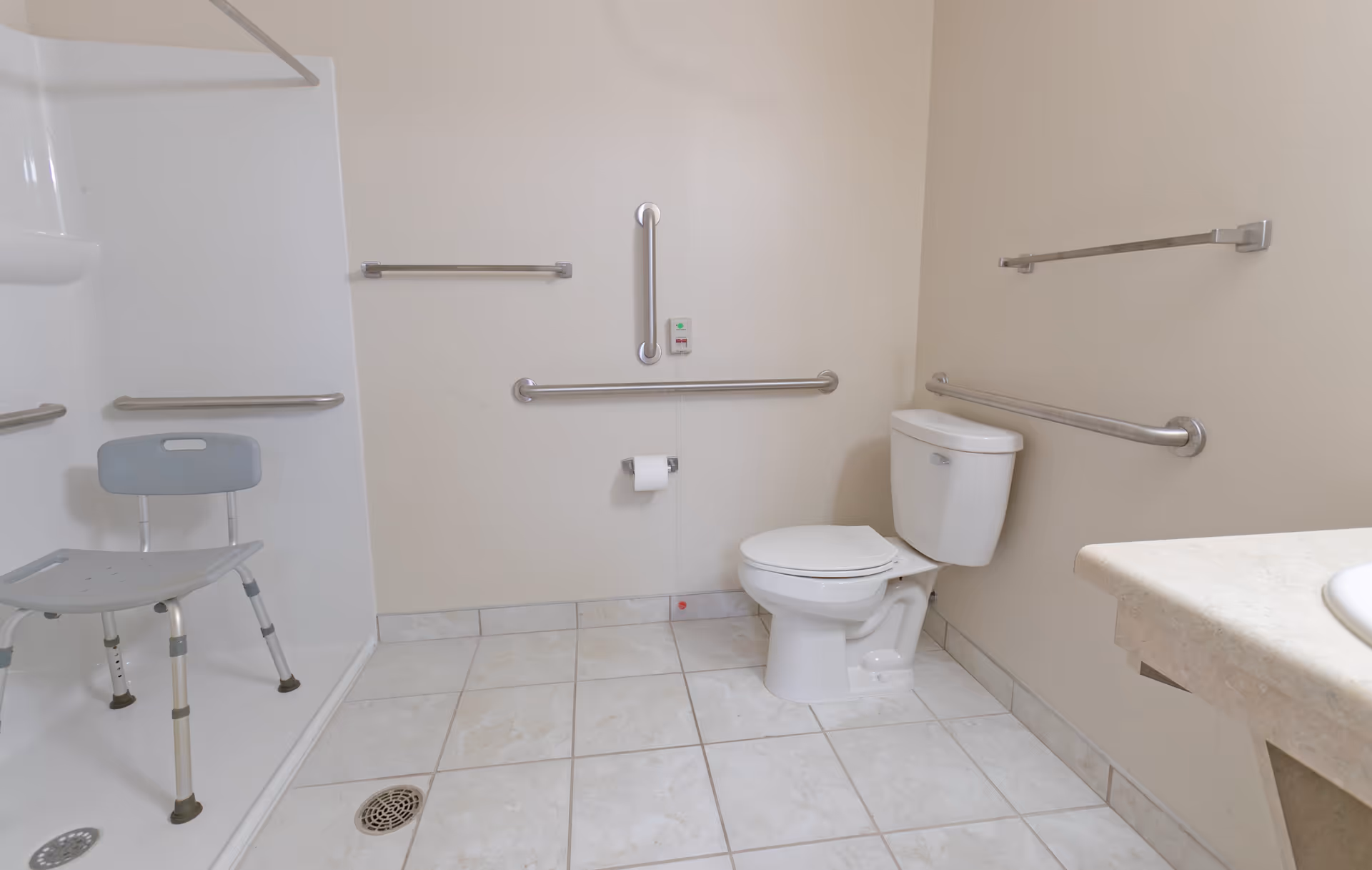 Accessible bathroom with a toilet, multiple grab bars, a shower with a shower chair, and a sink.