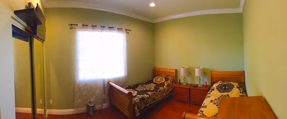 A small bedroom with two single wooden beds, each with patterned blankets. Between the beds is a wooden nightstand with two lamps and some small items. The walls are painted light green, and there is a window with sheer white curtains letting in natural light. A mirrored closet door is visible on the left side of the room.