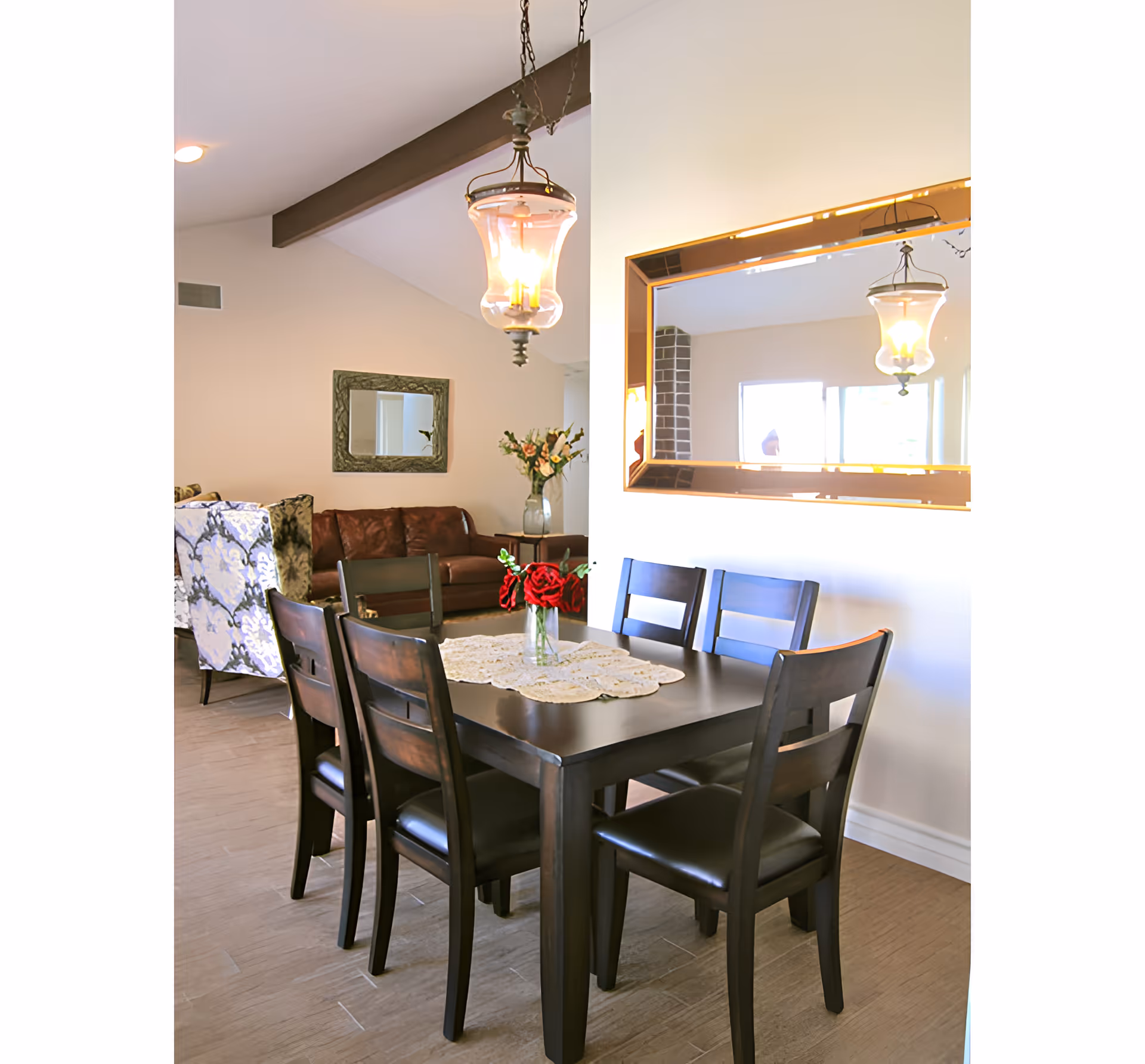 A dining area with a dark wooden table and six matching chairs. A vase with red flowers is placed on a lace doily in the center of the table. A large rectangular mirror with a gold frame hangs on the wall, reflecting a hanging light fixture. In the background, there is a living room area with a brown leather sofa, a floral patterned armchair, a wall mirror, and a vase with flowers on a side table.