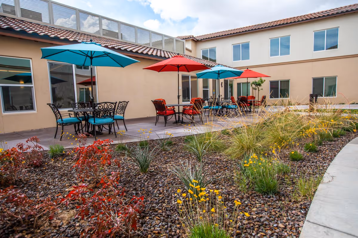 Outdoor patio area with tables and chairs under red and blue umbrellas, surrounded by a landscaped garden with rocks, grass, and yellow flowers, adjacent to a two-story building with multiple windows.