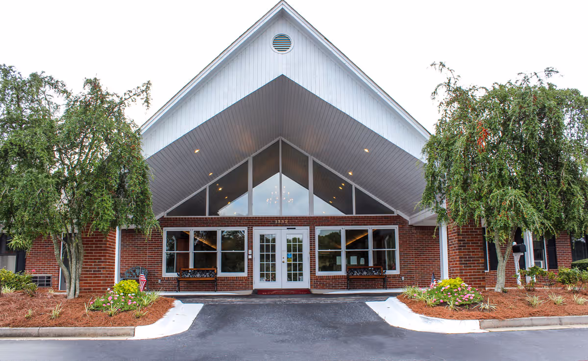 Front exterior view of a brick building with a large peaked roof and glass entrance doors. There are two benches on either side of the entrance and landscaped areas with small trees and flowers in front.