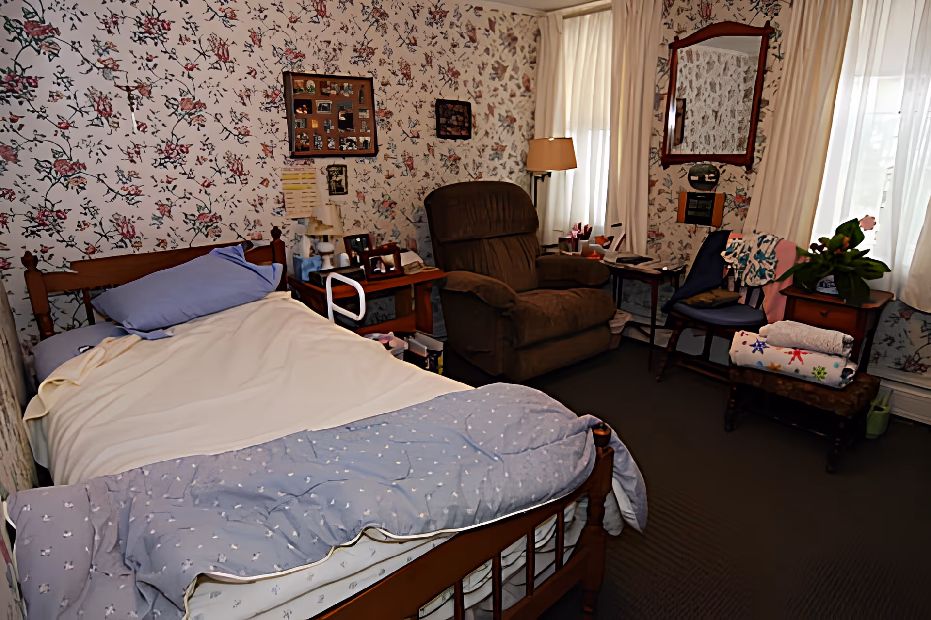 A cozy bedroom with floral wallpaper, a wooden bed with light-colored bedding, a brown recliner chair, a small wooden side table with framed photos, a lamp, and a chair with folded blankets and a potted plant. Light filters in through sheer curtains on two windows.