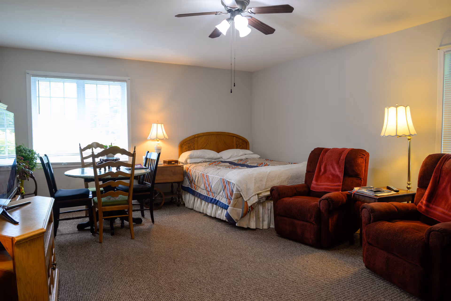 A cozy room with a bed against the wall, two red armchairs with red throws, a small wooden dining table with four chairs near a window, a ceiling fan with lights, and two lamps providing warm lighting.