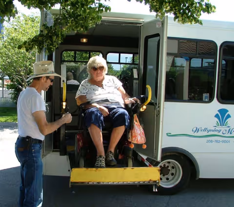 An elderly woman in a wheelchair is being assisted by a man wearing a hat as she is lifted on a wheelchair lift into a white shuttle bus with the logo and name Wellspring Meadows Assisted Living on the side.