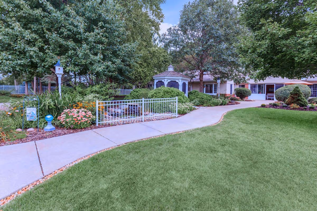 A landscaped outdoor garden area with a paved walkway, green grass, various bushes, flowers, and trees. There is a white gazebo in the background near a building with large windows.